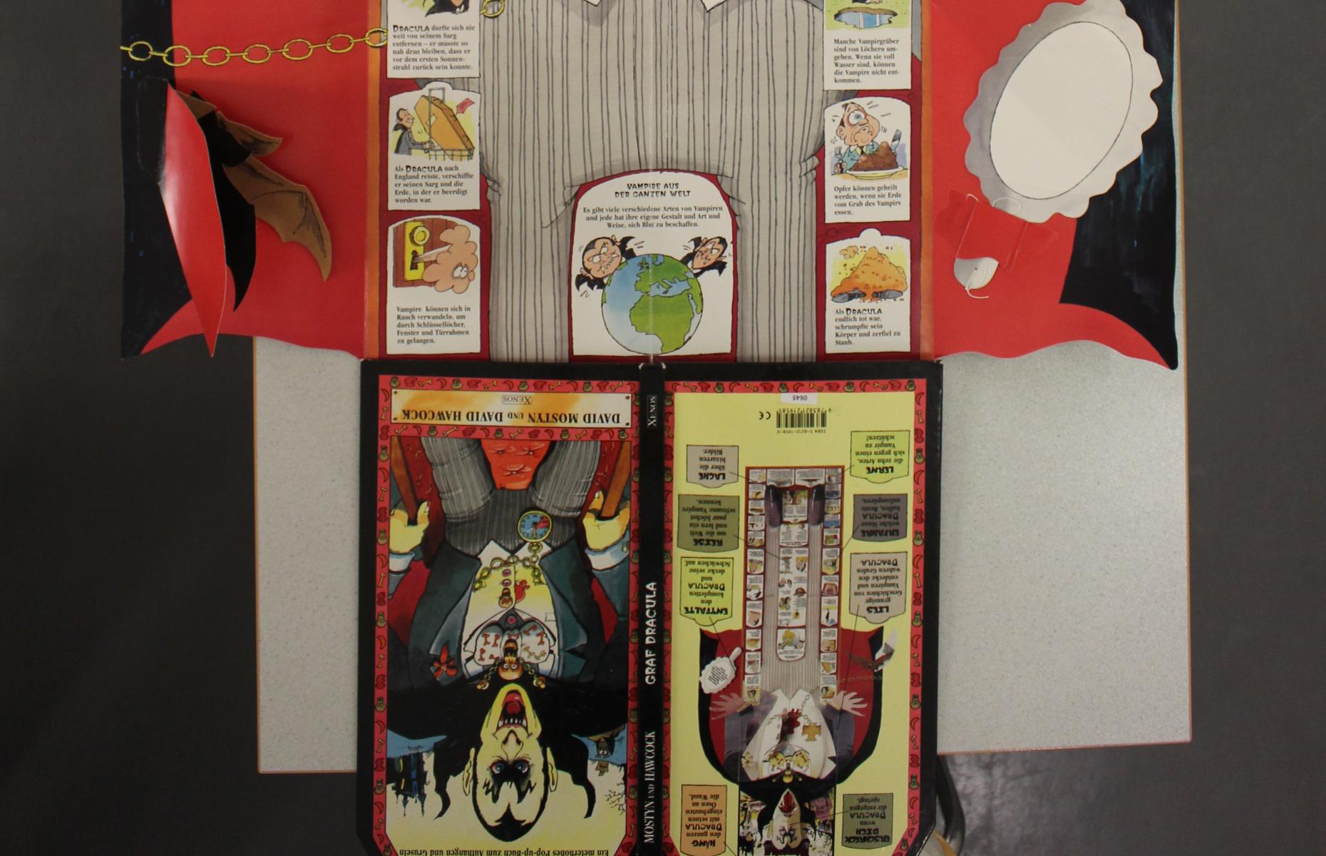 Unterer Teil des vollständig aufgeklappten Pop-up-Buch "Graf Dracula". Zu sehen sind der untere Teil von Graf Draculas Umhang, die Texte des Buches sowie die Vorder- und Rückseite des Buches.