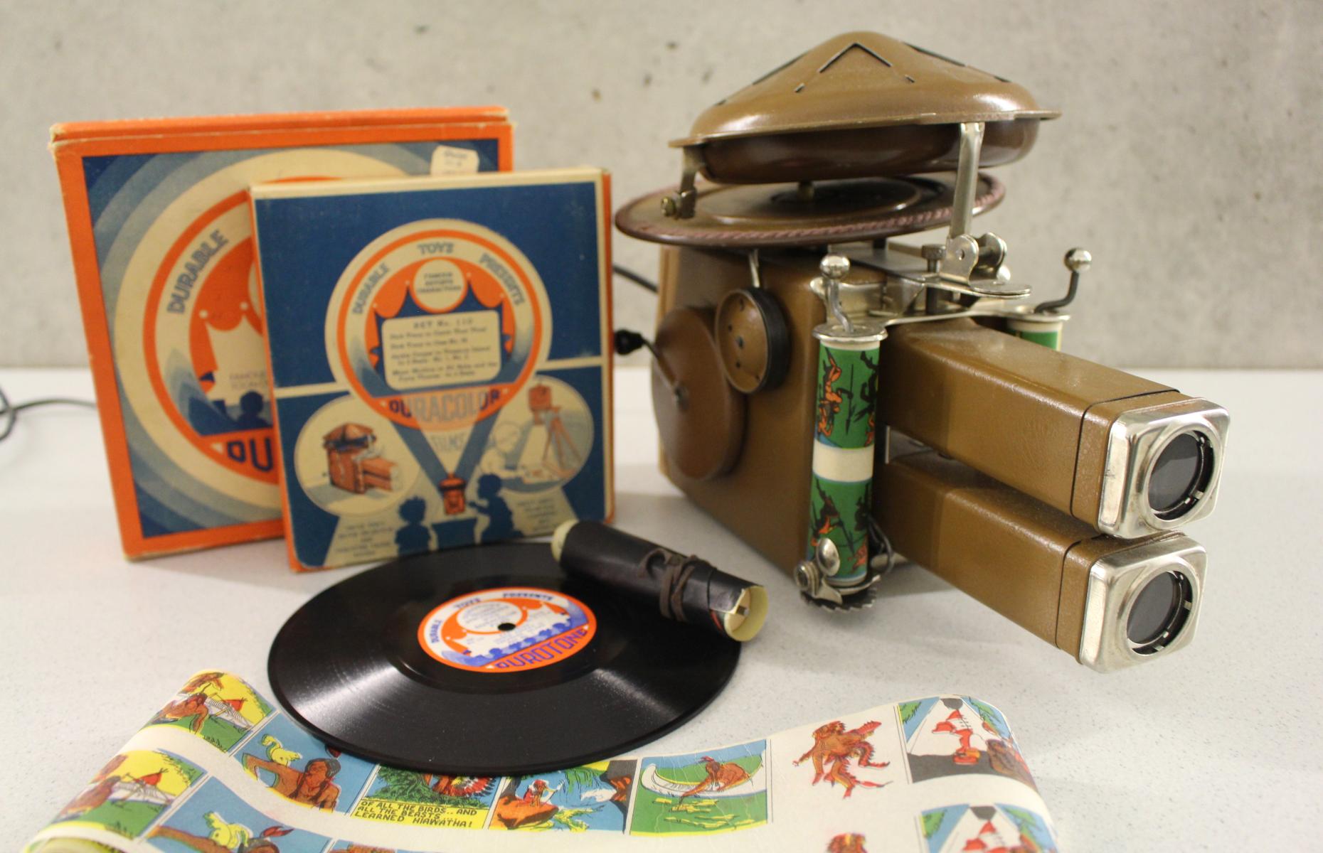 Der Projektionsapparat mit den Aufbewahrungsschachteln für die Filmrollen, einer Schallplatte und einem aufgerolltem Film.